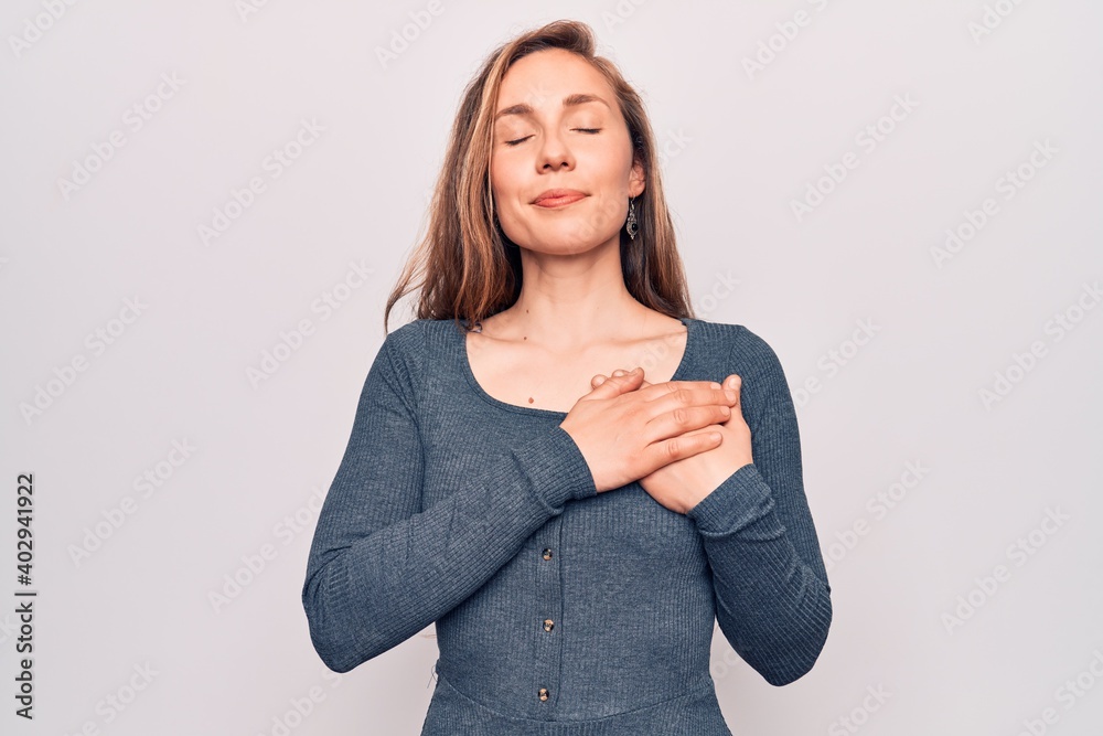 Young beautiful blonde woman standing over white isolated background smiling with hands on chest, eyes closed with grateful gesture on face. health concept. Young beautiful blonde woman standing over white isolated background smiling with hands on chest, eyes closed with grateful gesture on face. health concept.