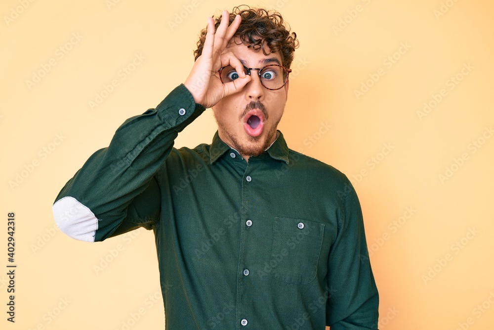 Young caucasian man with curly hair wearing casual clothes and glasses doing ok gesture shocked with surprised face, eye looking through fingers. unbelieving expression.