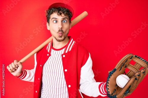 Young caucasian man with curly hair wearing baseball uniform holding golve and bat making fish face with mouth and squinting eyes, crazy and comical. photo