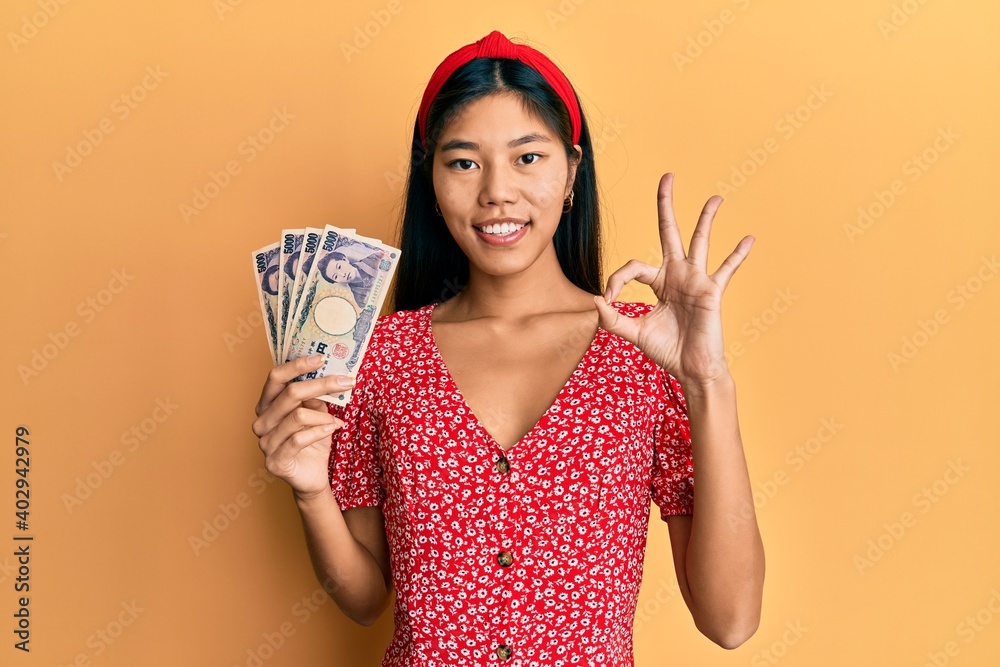 Young chinese woman holding 5000 japanese yen banknotes doing ok sign with fingers, smiling friendly gesturing excellent symbol