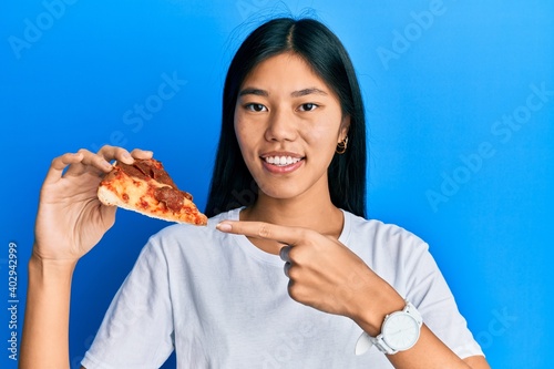 Young chinese woman eating tasty pepperoni pizza smiling happy pointing with hand and finger