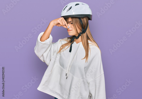 Teenager caucasian girl wearing bike helmet smelling something stinky and disgusting, intolerable smell, holding breath with fingers on nose. bad smell