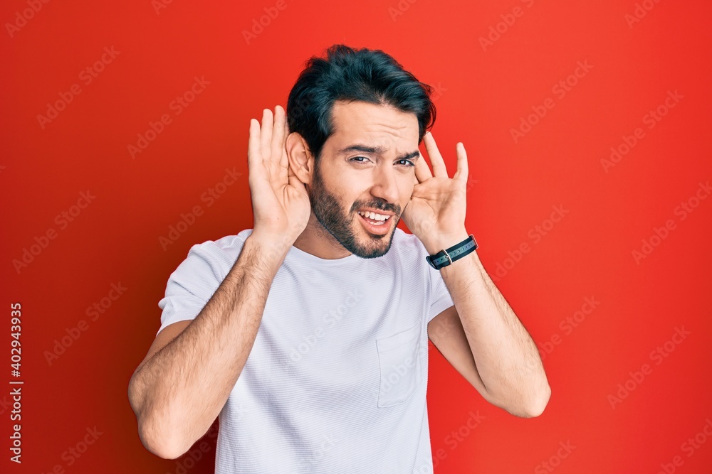 Fototapeta premium Young hispanic man wearing casual white tshirt trying to hear both hands on ear gesture, curious for gossip. hearing problem, deaf