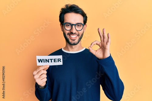 Young hispanic man wearing glasses holding winter word on paper doing ok sign with fingers, smiling friendly gesturing excellent symbol © Krakenimages.com