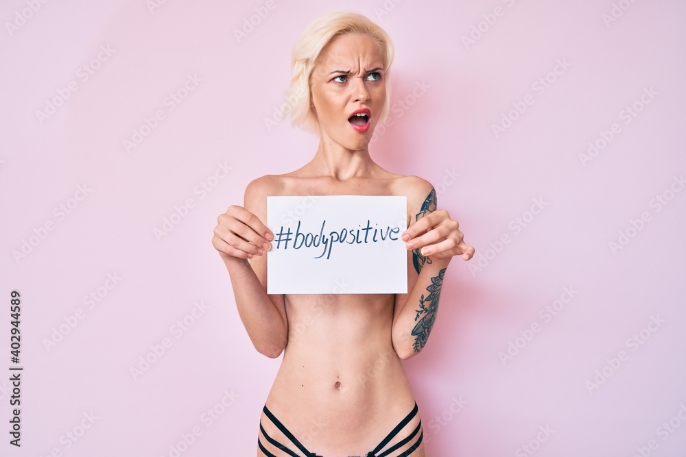 Young blonde woman with tattoo standing shirtless holding banner with body positive message angry and mad screaming frustrated and furious, shouting with anger. rage and aggressive concept. Young blonde woman with tattoo standing shirtless holding banner with body positive message angry and mad screaming frustrated and furious, shouting with anger. rage and aggressive concept.
