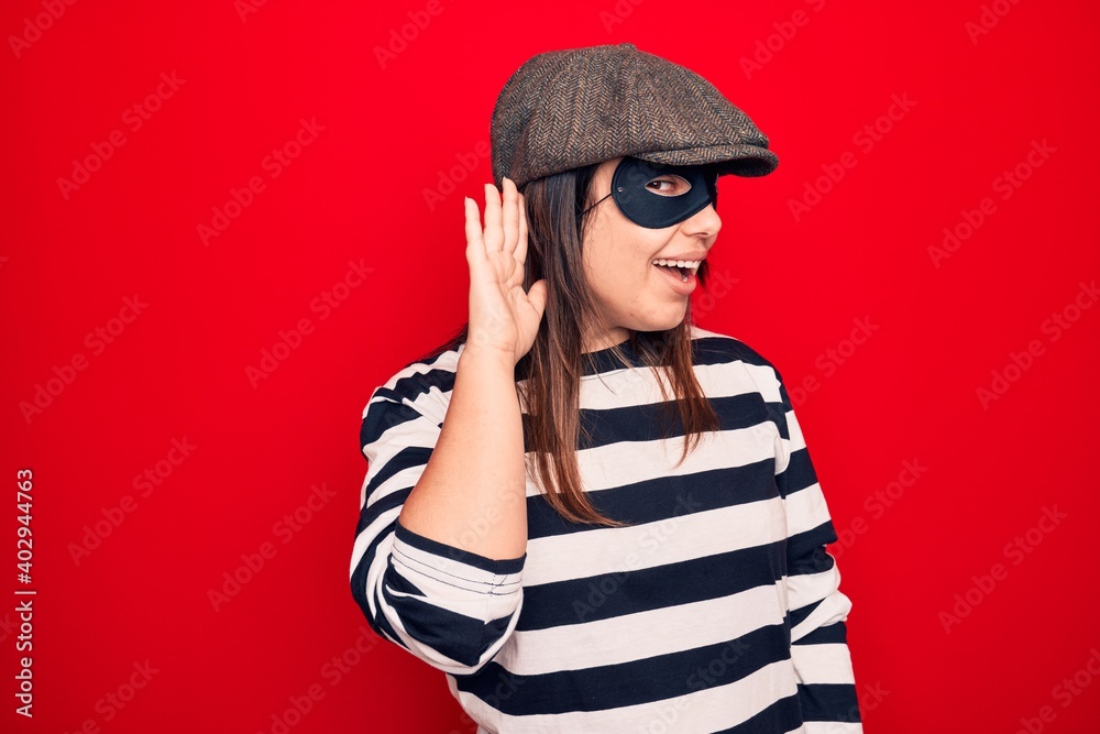 Young beautiful brunette burglar woman wearing cap and mask over isolated red background smiling with hand over ear listening and hearing to rumor or gossip. Deafness concept. Young beautiful brunette burglar woman wearing cap and mask over isolated red background smiling with hand over ear listening and hearing to rumor or gossip. Deafness concept.