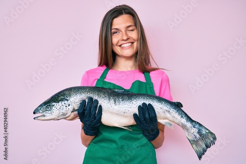 Beautiful caucasian woman fishmonger selling fresh raw salmon winking looking at the camera with sexy expression, cheerful and happy face.