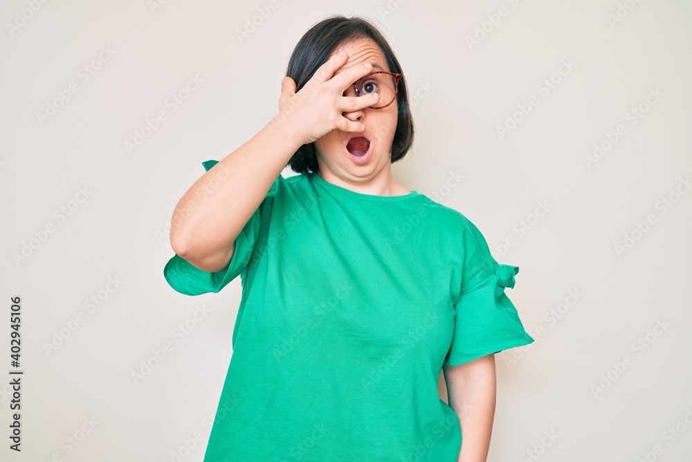Brunette woman with down syndrome wearing casual clothes and glasses peeking in shock covering face and eyes with hand, looking through fingers afraid Brunette woman with down syndrome wearing casual clothes and glasses peeking in shock covering face and eyes with hand, looking through fingers afraid