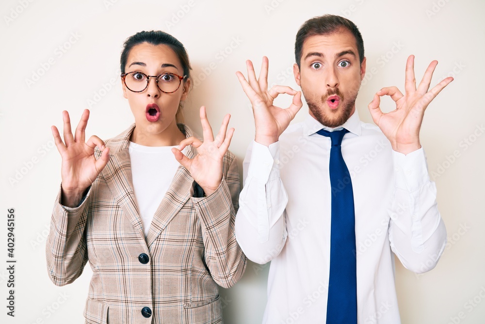 Beautiful couple wearing business clothes looking surprised and shocked doing ok approval symbol with fingers. crazy expression Beautiful couple wearing business clothes looking surprised and shocked doing ok approval symbol with fingers. crazy expression