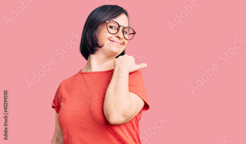 Brunette woman with down syndrome wearing casual clothes and glasses smiling with happy face looking and pointing to the side with thumb up. photo