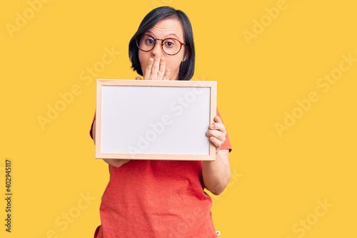 Brunette woman with down syndrome holding empty white chalkboard covering mouth with hand, shocked and afraid for mistake. surprised expression