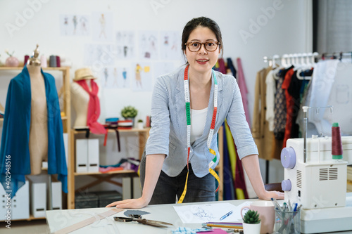 Successful fashion designer woman worker in studio. attractive young asian chinese female staff wear glasses putting hands on work desk and looking at camera with smile while standing in workshop.