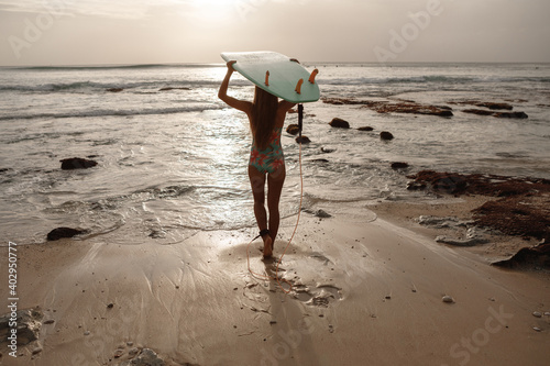  Attractive young woman in surf swimsuit is standing on beach with blue surfboard in hands over her head, look at sunse and go to the ocean catch waves
