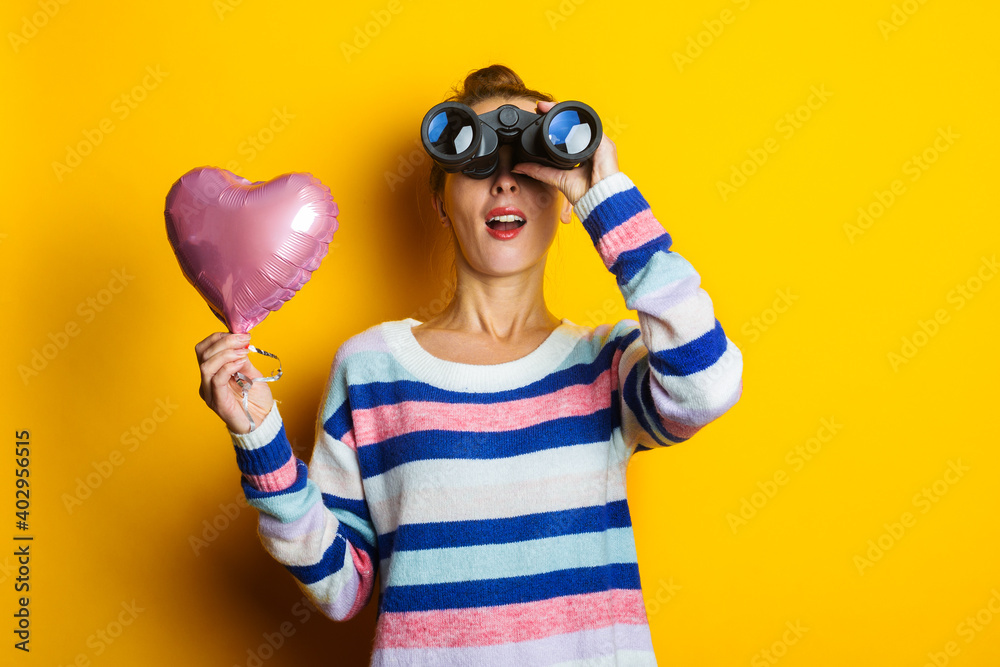 Young woman in a sweater holds an air balloon heart and looks through binoculars on a yellow background. Valentine's day composition