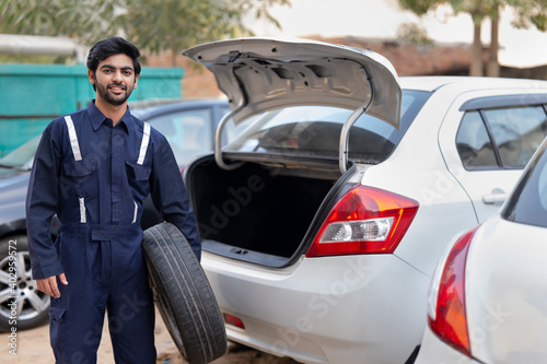 A YOUNG MECHANIC HOLDING TYRE STANDING NEAR A CAR AND SMILING AT CAMERA 