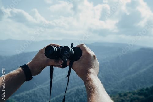 Hands holding binoculars on mountains forest nature background, looking through binoculars, travel, search and search concept.