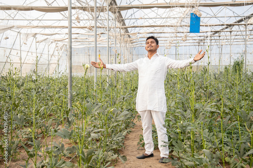Young happy indian farmer standing with open arms at his poly house or greenhouse, agriculture business and rural prosperity concept. man wearing white kurta pajama cloths, copy space.