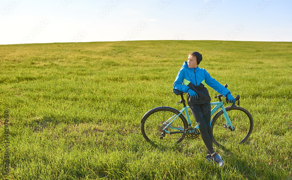 A young cyclist in a tracksuit stopped in a field on a background of green grass in sunny weather.