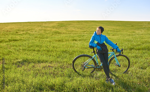A young cyclist in a tracksuit stopped in a field on a background of green grass in sunny weather.