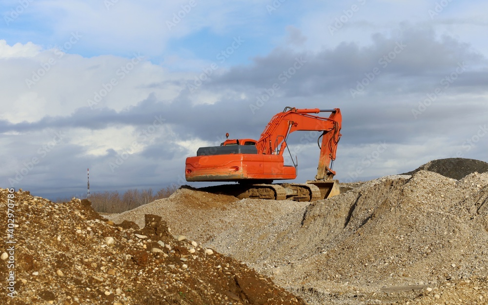 Excavator on large heaps of gravel and dirt. Cloudy sky on background.