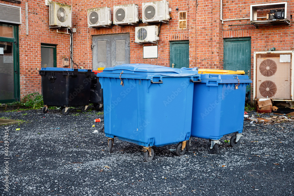 Back yard of an office building with colored rubbish bins, fire doors and utility room entrance. Many air conditioner units mounted on a brick wall. Dirt on the ground Back yard of an office building with colored rubbish bins, fire doors and utility room entrance. Many air conditioner units mounted on a brick wall. Dirt on the ground