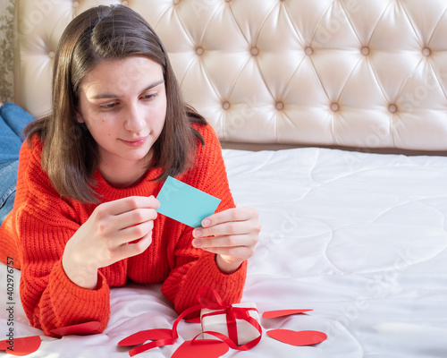 Young caucasian girl lying on the bed reading a message card and looking at a gift with heart shapes, copy space. For use on Valentine's Day, Birthday