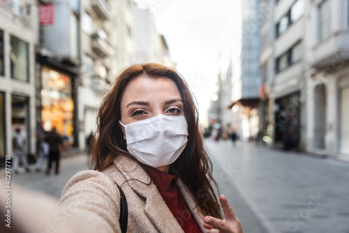 Beautiful girl wearing protective medical mask and fashionable clothes takes selfie with a smart phone at street. New normal lifestyle concept. © epic_images