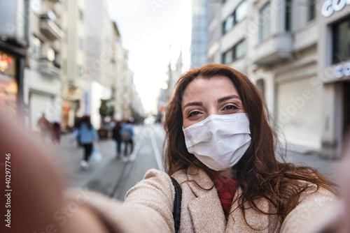 Beautiful girl wearing protective medical mask and fashionable clothes takes selfie with a smart phone at street. New normal lifestyle concept.