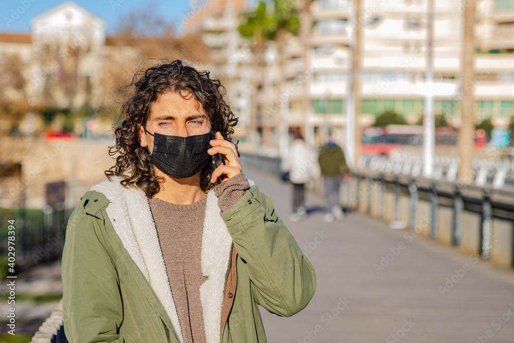 Young Curly Long Hair Man With Mask and Green Jacket Talking to the Phone with Disgusted Face Expression Hearing Bad News at the Bridge