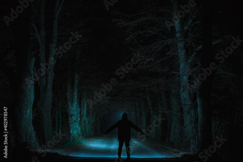 A deserted path with a standing figure in the middle between dense trees on the sides at night illuminated by lights to the center.