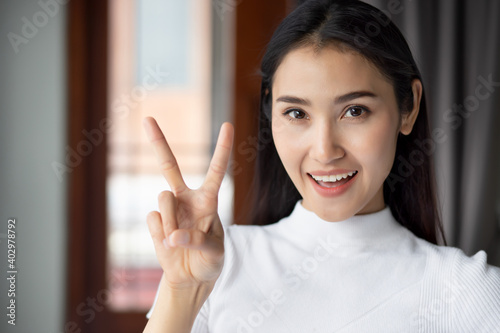 asian woman pointing two fingers V for victory sign, portrait of happy smiling asian woman pointing up 2 fingers for number two or 2 points concept by indoor casual Chinese asian adult woman model