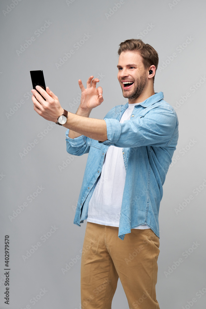 Smiling young man wearing jeans shirt taking selfie photo on smartphone or making video call standing over grey studio background Smiling young man wearing jeans shirt taking selfie photo on smartphone or making video call standing over grey studio background