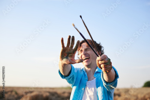 Male artist with dirty with paint hands, holding brushes in front of his face smiling. Artistic education activity. Painting workshop outdoors on wheat field. Summer leisure entertainment.