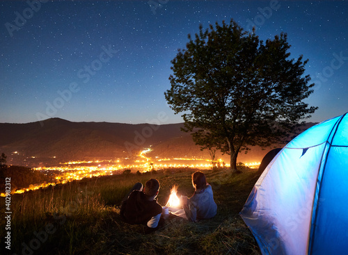 Back view of young couple tourists resting at campfire beside camp and blue tent, drinking coffee, enjoying amazing night sky full of stars. On background starry sky, mountains, tree and luminous town