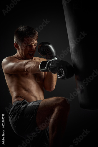 muscular boxer in black boxing gloves punching in boxing bag on dark background