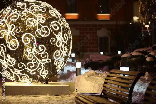 An empty wooden bench on a snowy background of snowdrifts and a huge glowing Christmas ball with a garland and an ornate pattern.Festive decoration for the New Year in the city park