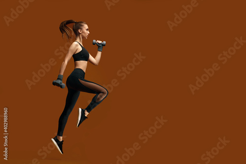 athletic beautiful woman in black sportswear training with dumbbells isolated on yellow background. girl jumping with dumbbells. sport and fitness concept. copy space