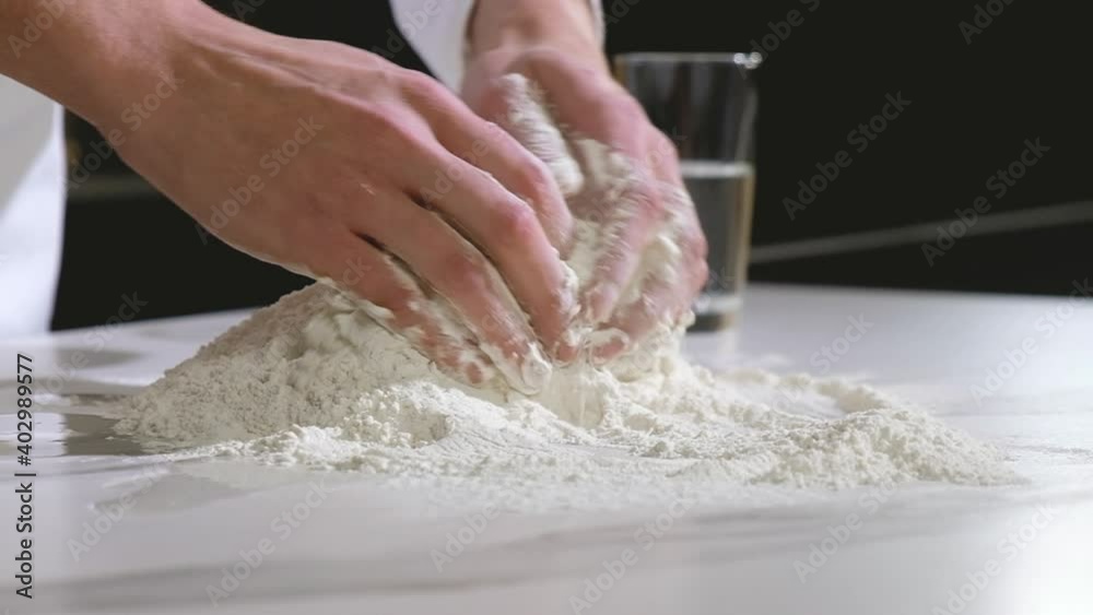 Professional baker works in kitchen of bakery shop. cook carefully mixes white wheat flour with chicken egg on table with his hands. Kneading dough. Preparation of dough for baking bread and pizza. Professional baker works in kitchen of bakery shop. cook carefully mixes white wheat flour with chicken egg on table with his hands. Kneading dough. Preparation of dough for baking bread and pizza.