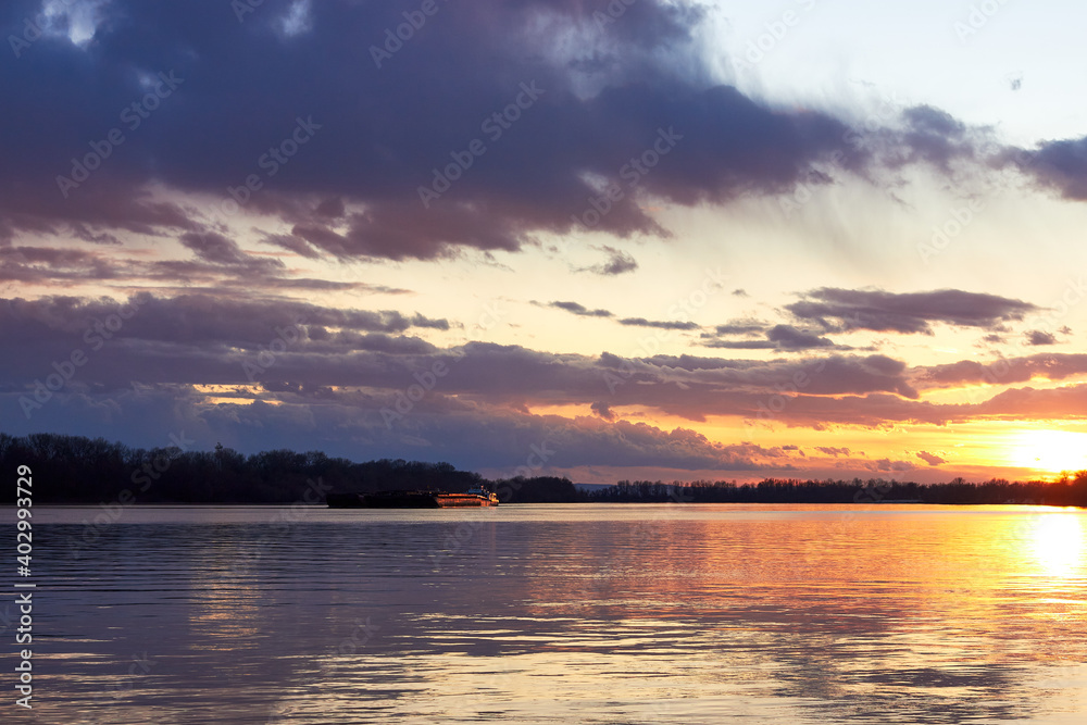 Landscape view of the river in the evening time, with wonderful blue cloudy sky during the yellow sunset lighting and a barge (ship) on the horizon