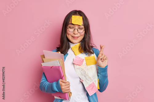 Positive schoolgirl crosses fingers believes in good luck on exam wears round spectacles stuck with papers and sticky notes written information to remember makes crib. Student uses cheat sheets.
