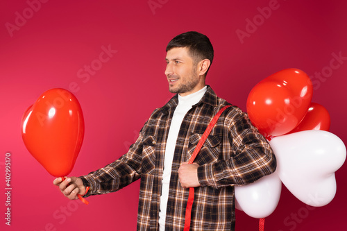Profile photo. A man with stretches out a red heart balloon holding a bunch of balloons on his shoulder. Red background. © Viorel