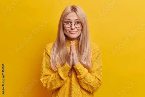 Pleased light haired European woman looks hopefully keeps palms in pray gesture asks for help dressed in casual jumper isolated over vivid yellow background. People and body language concept © wayhome.studio 