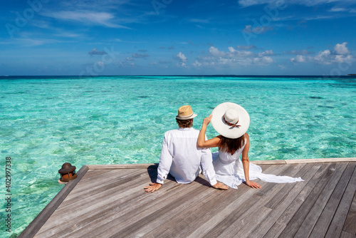 A traveler couple with hats sits together on a wooden pier and enjoys the view to the turquoise ocean of the Maldives islands