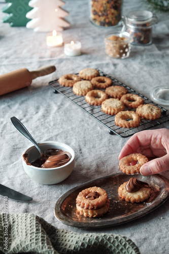 Christmas Linzer cookies filled on cooling rack, metal plate. Sandwich cookies filled with hazelnut spread, nougat cream. Hand holds cookie. Xmas trees and candles on beige linen tablecloth.