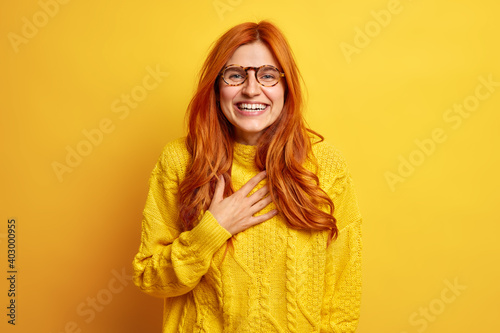 Portrait of overjoyed redhead woman smiles broadly keeps hand on chest smiles broadly expresses good emotions feels pleased wears transparent glasses casual sweater isolated over yellow background © wayhome.studio 