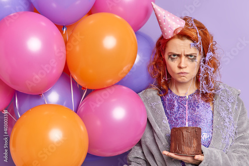Insulted woman looks angrily from under forehead expresses negative emotions being offended on her birthday has broken heart wears cone party hat dressing gown holds bunch of inflated balloons