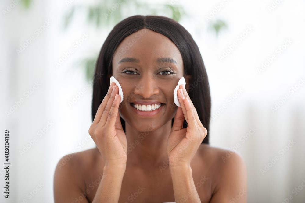 Smiling young african american woman doing daily morning skincare procedures, cleans face skin with natural lotion