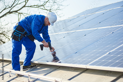 Male engineer in blue suit and protective helmet installing stand-alone solar photovoltaic panel system using screwdriver. Electrician mounting blue solar module on roof of modern house