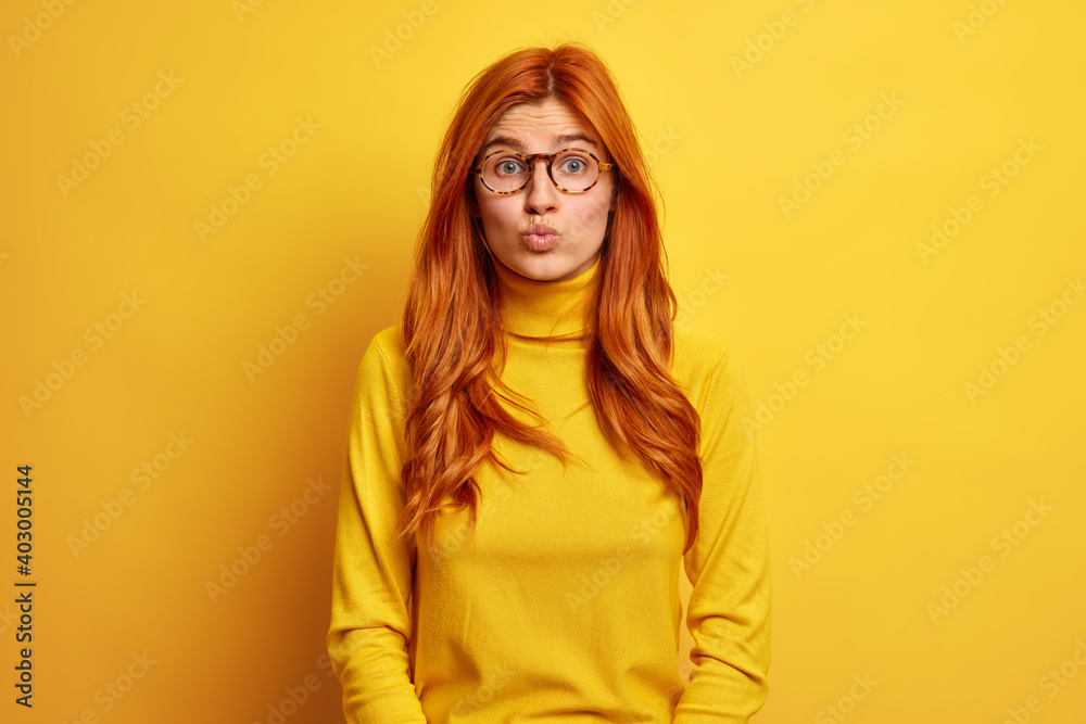 Portrait of pleasant looking redhead woman keeps lips rounded wants to kiss someone has romantic look wears optical glasses turtleneck isolated over yellow background. Human face expressions
