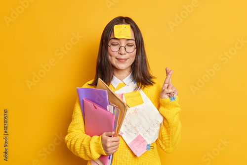 Pleased Asain female student believes in good luck at exam stands with eyes closed and fingers crossed believes dreams come true stuck with papers holds folders isolated over yellow background